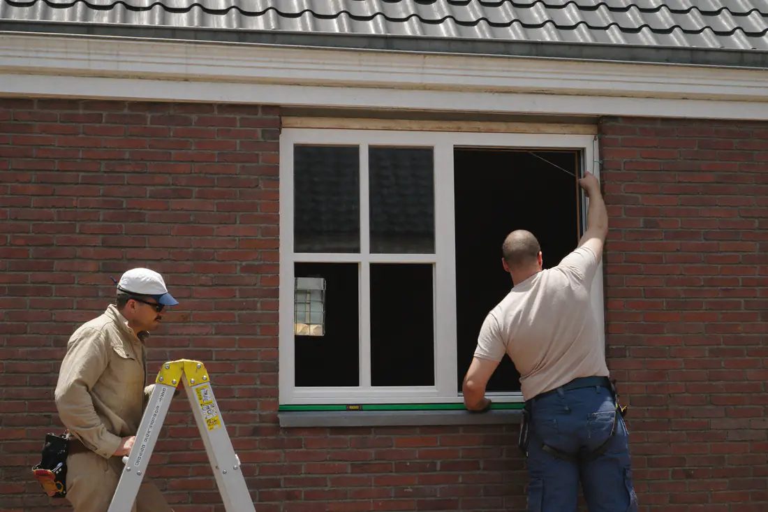 Twee monteurs plaatsen een nieuw wit kozijn in de gevelopening van een bakstenen huis